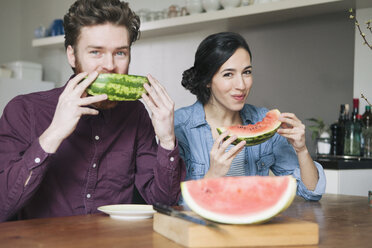 Porträt eines glücklichen jungen Paares, das am Küchentisch Wassermelone isst - FSIF00872