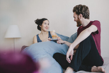 Happy young woman holding birthday cupcake with candle while looking at man on bed - FSIF00879