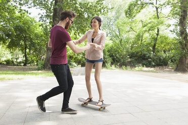 Junger Mann, der einer Frau beim Skateboardfahren auf dem Gehweg im Park hilft - FSIF00964