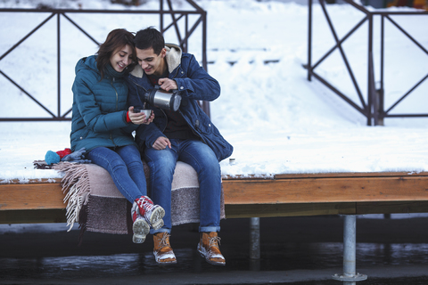 Junger Mann serviert seiner Freundin auf dem Pier sitzend Kaffee in voller Länge, lizenzfreies Stockfoto