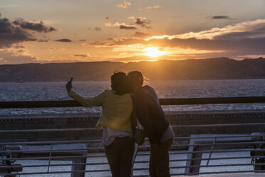 Rückansicht eines Paares, das bei Sonnenuntergang ein Selfie an der Reling am Meer macht - FSIF00997