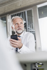 Älterer Geschäftsmann mit Handy am Schreibtisch im Büro denkt - UUF12732