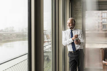 Smiling mature businessman standing at the window looking out - UUF12740