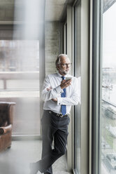 Mature businessman standing at the window holding cell phone - UUF12742