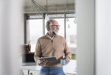 Smiling casual mature businessman holding tablet and cup of coffee in conference room in office - UUF12768