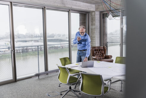 Älterer Geschäftsmann mit Laptop und Smartphone im Konferenzraum im Büro - UUF12802