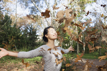 Trockene Blätter, die auf eine glückliche Frau fallen, die mit ausgestreckten Armen in einem Park im Herbst steht - FSIF01044