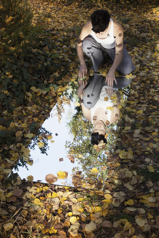 Hoher Blickwinkel auf eine Frau und Bäume, die sich im Spiegel spiegeln, während sie auf trockenen Blättern im Park sitzen, lizenzfreies Stockfoto