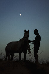 Silhouette Mann stehend mit Pferd auf Feld gegen blauen Himmel in der Abenddämmerung - FSIF01052