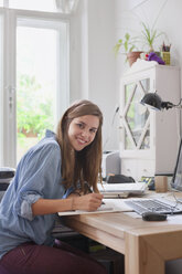 Porträt einer lächelnden jungen Frau, die an einem Computertisch in einem Zimmer schreibt - FSIF01068