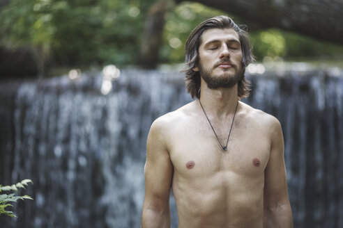 Hemdloser junger Mann mit geschlossenen Augen steht gegen Wasserfall im Wald - FSIF01106