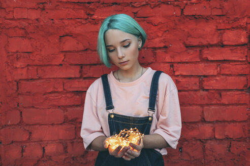 Teenage girl holding illuminated string lights while standing against brick wall - FSIF01116