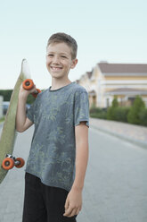 Porträt eines lächelnden Jungen, der ein Skateboard hält und auf der Straße steht - FSIF01125