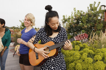 Junge Frau spielt Gitarre auf der Terrasse, während Freundinnen im Hintergrund stehen - FSIF01177