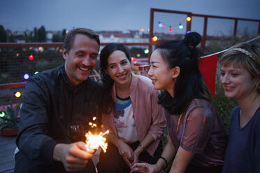 Happy man holding sparkler while sitting with female friends at night - FSIF01184