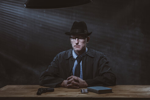 Porträt eines jungen Mannes, der mit einer Pistole am Tisch sitzt, vor einer schwarzen Wand - FSIF01232