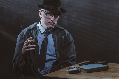 Young man sitting at table while smoking cigarette against black wall - FSIF01234