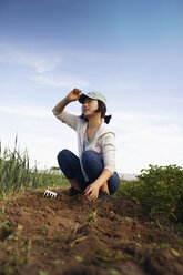 Hockende Frau, die nach oben schaut, während sie auf einem Feld auf einem Bauernhof an einem sonnigen Tag arbeitet - FSIF01275