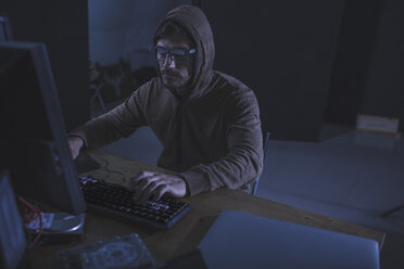 Serious Computer Hacker mit Kapuzen-Shirt mit Desktop-Computer am Tisch in verlassenen Raum - FSIF01284