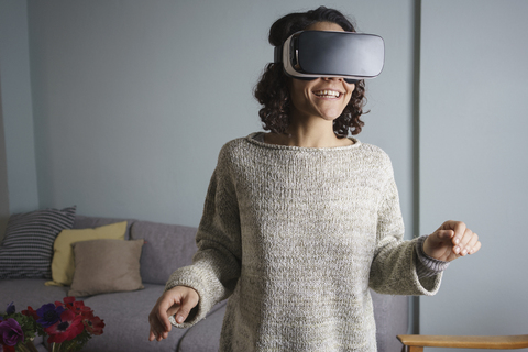 Happy woman using virtual reality headset while standing in living room stock photo