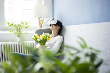 Lächelnde Frau sitzt auf der Couch und trägt eine VR-Brille und hält einen Strauß Tulpen - MOEF00805