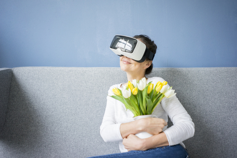 Lächelnde Frau sitzt auf der Couch und trägt eine VR-Brille und hält einen Strauß Tulpen, lizenzfreies Stockfoto