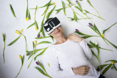 Lächelnde Frau mit VR-Brille auf dem Boden liegend inmitten von Tulpen, lizenzfreies Stockfoto