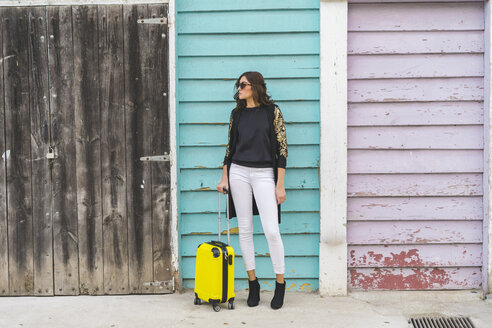 Junge Frau mit einer gelben Trolley-Tasche wartet vor einer Holzfassade - AFVF00059