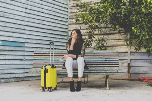 Young woman with yellow trolley bag waiting on a bench - AFVF00063