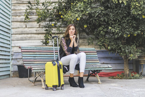 Junge Frau mit einer gelben Trolley-Tasche, die auf einer Bank wartet - AFVF00066