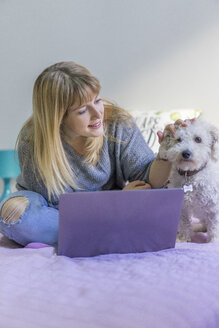 Frau mit Laptop sitzt auf dem Bett und streichelt ihren Hund - AFVF00075