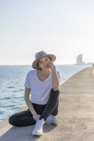 Spain, Barcelona, portrait of woman wearing hat sitting at waterfront promenade stock photo