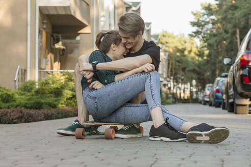 Glückliches junges Paar, das sich umarmt, während es auf einem Skateboard im Freien sitzt - FSIF01399