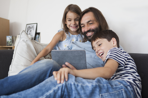 Glücklicher Vater mit Kindern, der auf dem Sofa zu Hause ein digitales Tablet benutzt, lizenzfreies Stockfoto