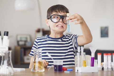 Junge mit schielenden Augen betrachtet Reagenzglas bei wissenschaftlichem Experiment im Haus, lizenzfreies Stockfoto