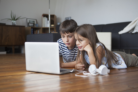 Geschwister benutzen Laptop, während sie zu Hause auf dem Hartholzboden liegen, lizenzfreies Stockfoto