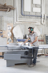 Männlicher Tischler in voller Länge bei der Arbeit mit einer Formatkreissäge in der Werkstatt - FSIF01444