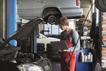 Mechaniker, der einen Laptop benutzt, während er den Motor eines Autos in der Werkstatt untersucht - FSIF01473