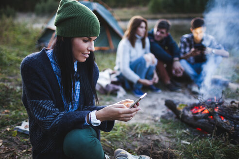 Junge Frau, die ein Mobiltelefon benutzt, während Freunde am Lagerfeuer auf einem Campingplatz sitzen - FSIF01499