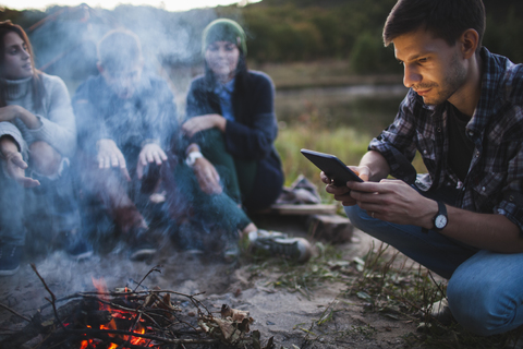 Junger Mann benutzt ein digitales Tablet, während er mit Freunden am Lagerfeuer sitzt, lizenzfreies Stockfoto