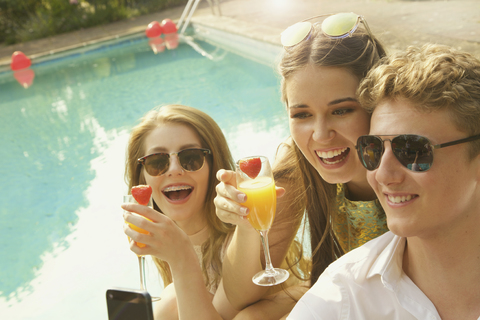 Fröhliche Frau mit Getränken in der Hand, während sie mit einem männlichen Freund am Pool sitzt, lizenzfreies Stockfoto