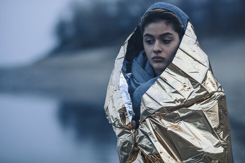 Nachdenkliches Teenager-Mädchen, eingewickelt in goldenes Plastik am Seeufer im Winter - FSIF01614