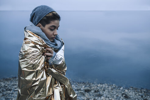 Teenager-Mädchen in goldenem Plastik eingewickelt am Seeufer im Winter - FSIF01619