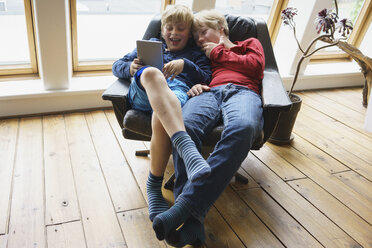 High angle view of happy brothers using digital tablet while resting on chair at home - FSIF01646