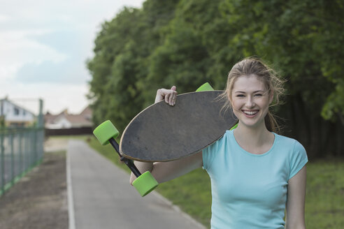 Porträt einer fröhlichen Frau mit Skateboard auf einem Fußweg im Park - FSIF01652