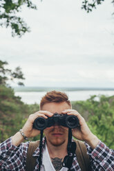 Nahaufnahme eines jungen Mannes mit Fernglas beim Wandern im Wald gegen den Himmel - FSIF01696