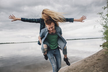 Happy man piggybacking girlfriend on lakeshore against sky - FSIF01701