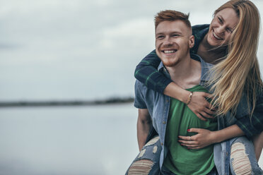 Happy man piggybacking cheerful girlfriend on lakeshore against sky - FSIF01709