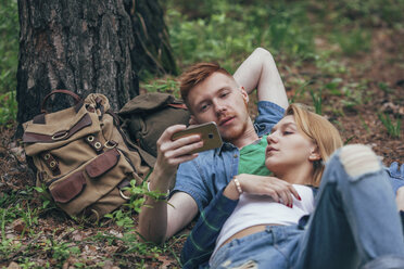 Junges Paar schaut in sein Smartphone, während es an einem Baum im Wald liegt - FSIF01714