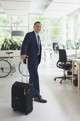 Confident businessman standing with suitcase at creative office - FSIF01769